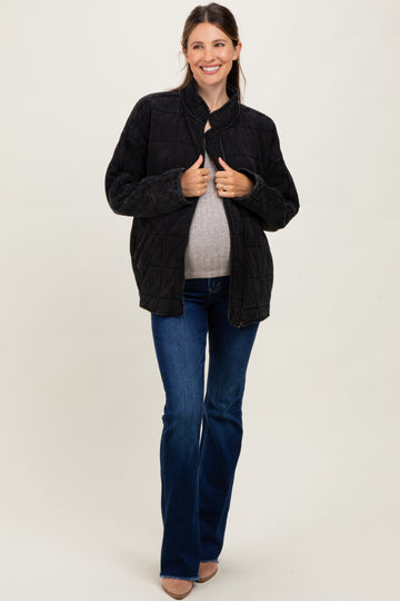 woman wearing a black quilted maternity jacket and a gray top, holding her baby bump while standing in flared jeans