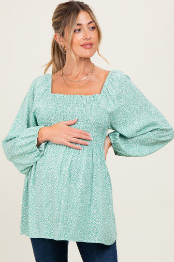 woman wearing a green floral maternity top with square neckline and puffed sleeves, gently resting her hands on her baby bump
