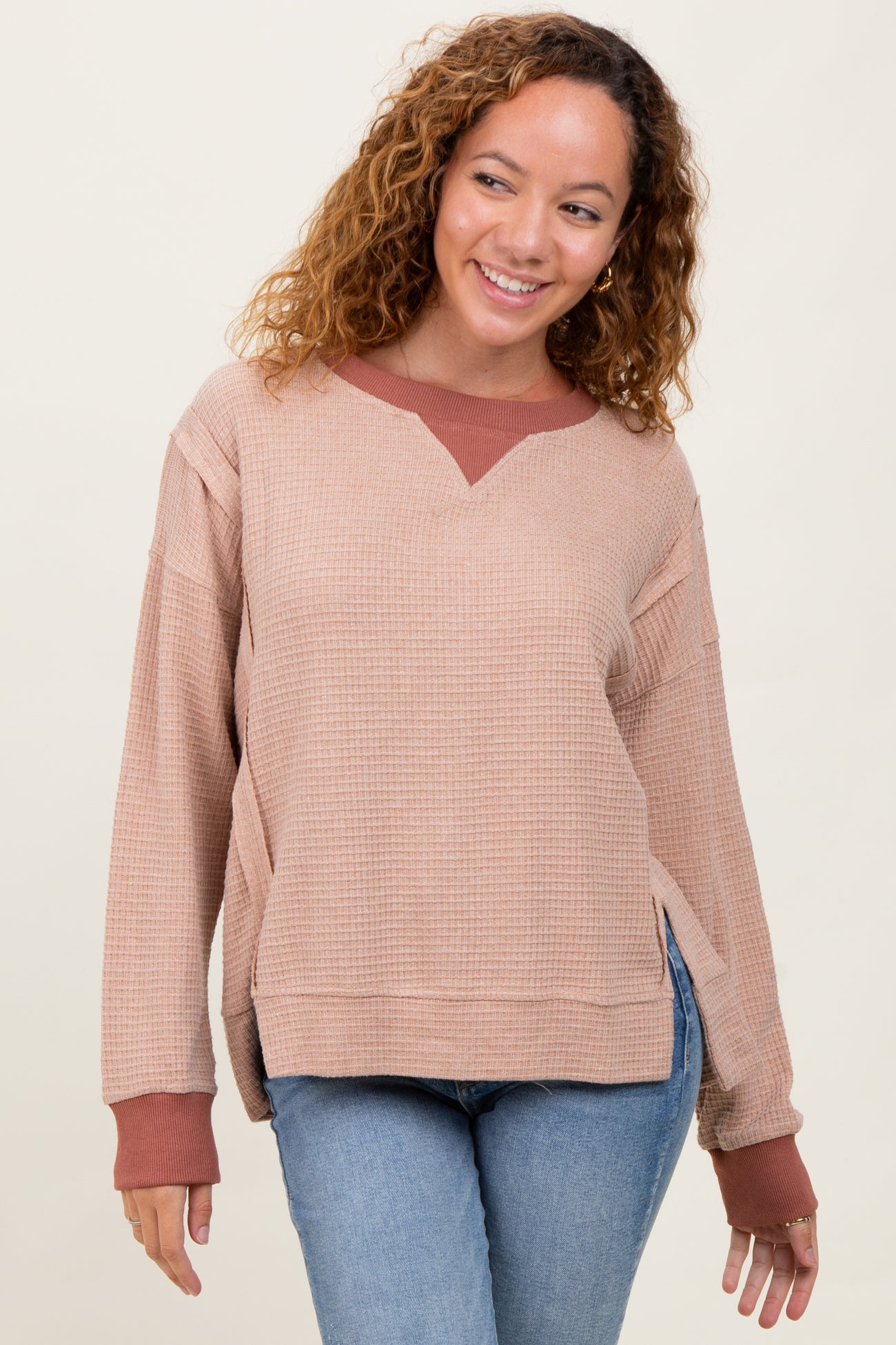 woman wearing a beige oversized waffle knit sweater with a maroon collar and side slits, paired with blue jeans