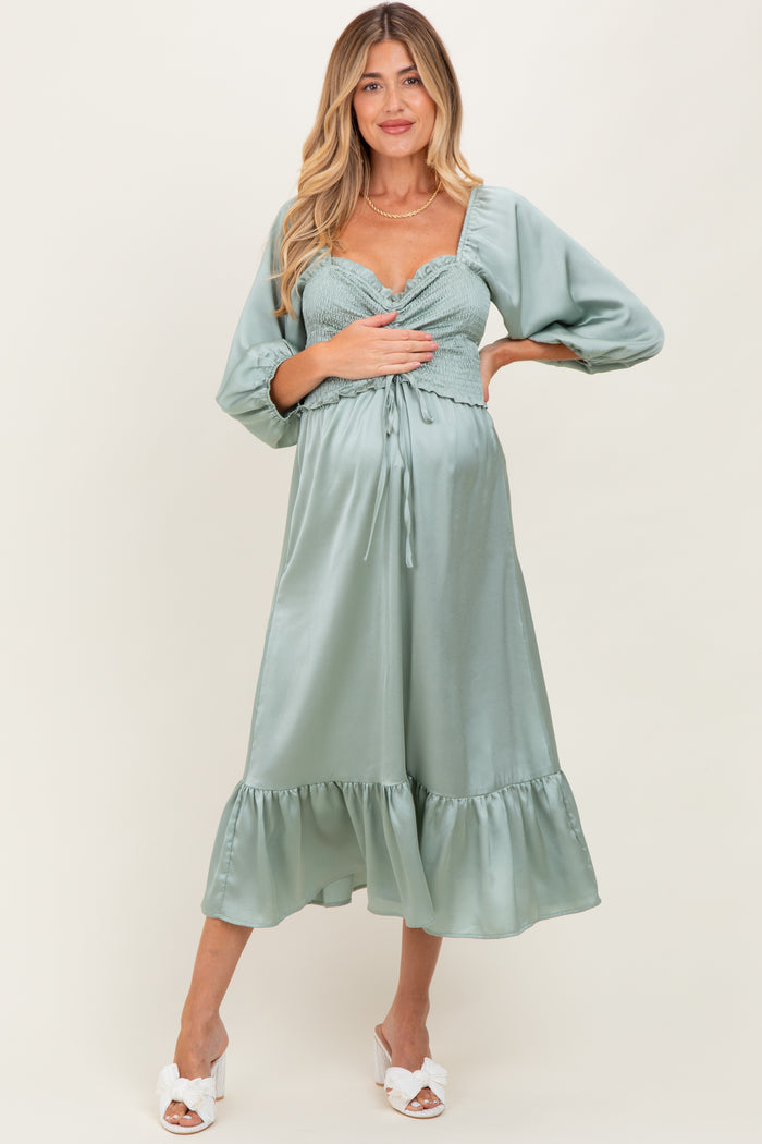 woman wearing a light green maternity dress with a smocked bodice and ruffled hem, holding her baby bump