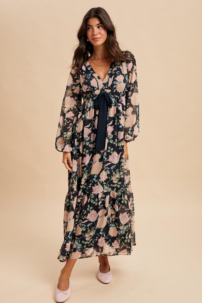 woman wearing a black floral maxi dress with long sleeves and a tie waist, standing on a neutral background