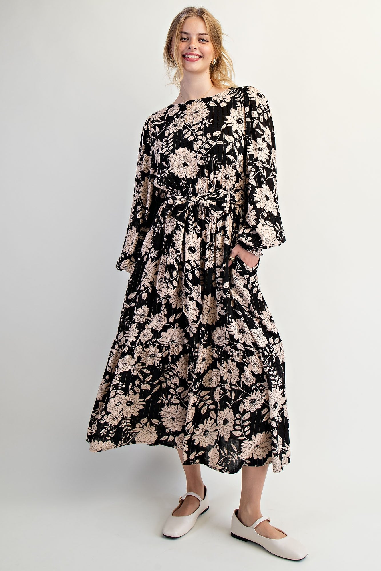 woman wearing a black floral maxi dress with long sleeves and pockets, smiling at the camera