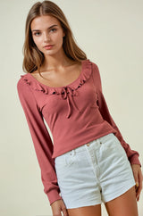 woman wearing a dusty rose long sleeve top with ruffle details and a tie at the neckline, paired with white shorts
