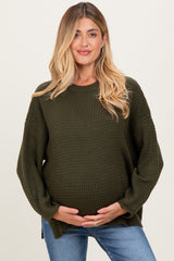 woman wearing a dark green oversized maternity sweater, holding her baby bump and paired with light blue jeans