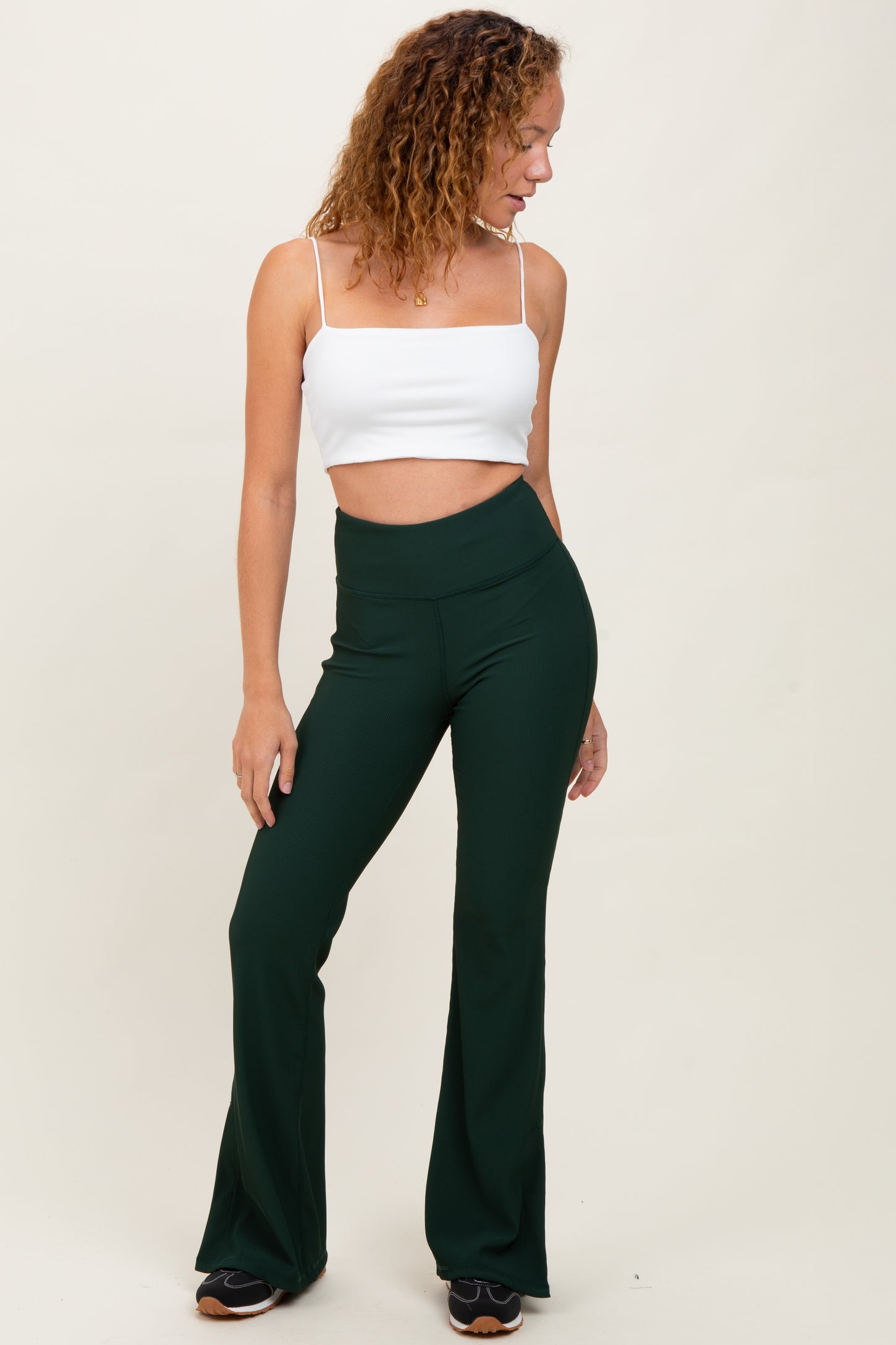 woman wearing a white cropped tank top and green flared pants, standing sideways with one hand on her hip