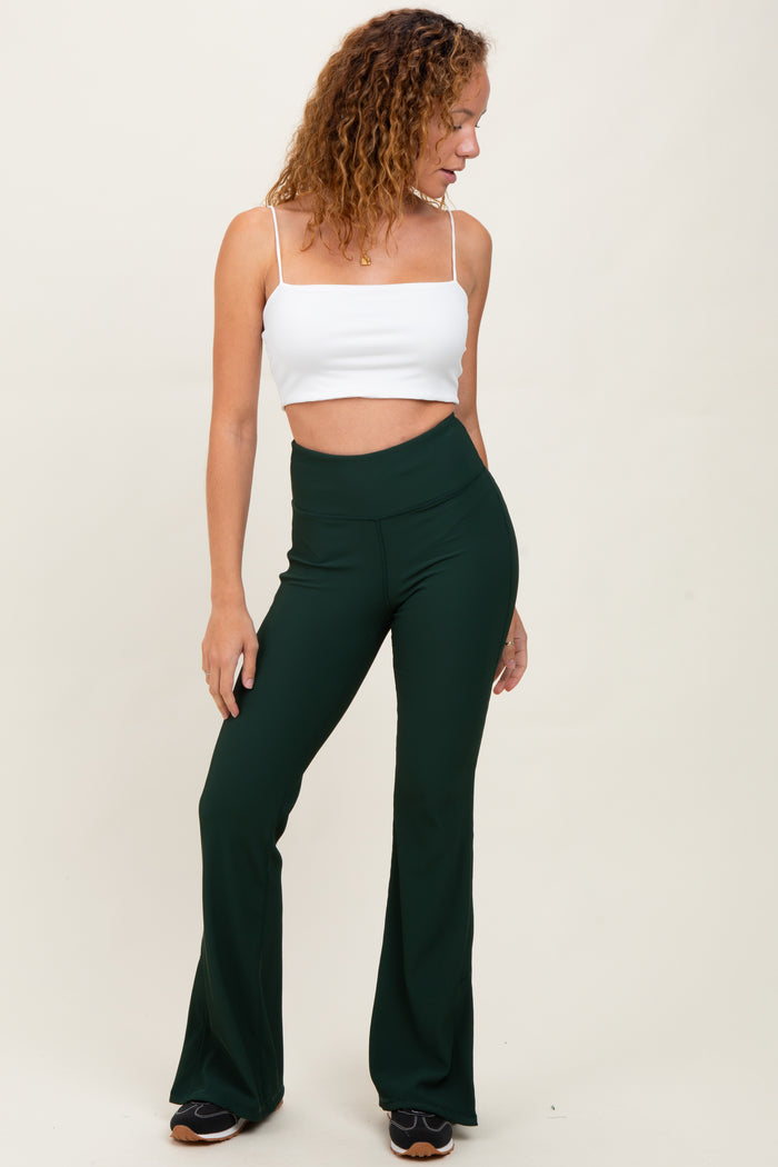 woman wearing a white cropped tank top and green flared pants, standing sideways with one hand on her hip