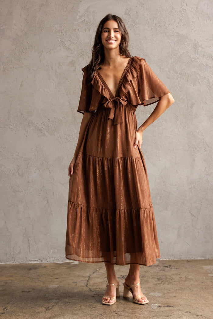 woman wearing a brown tiered maxi dress with ruffled sleeves and a tie waist, standing against a textured wall
