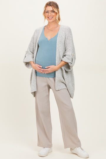 woman wearing a gray open cardigan, blue maternity top, and beige wide-leg pants, holding her baby bump