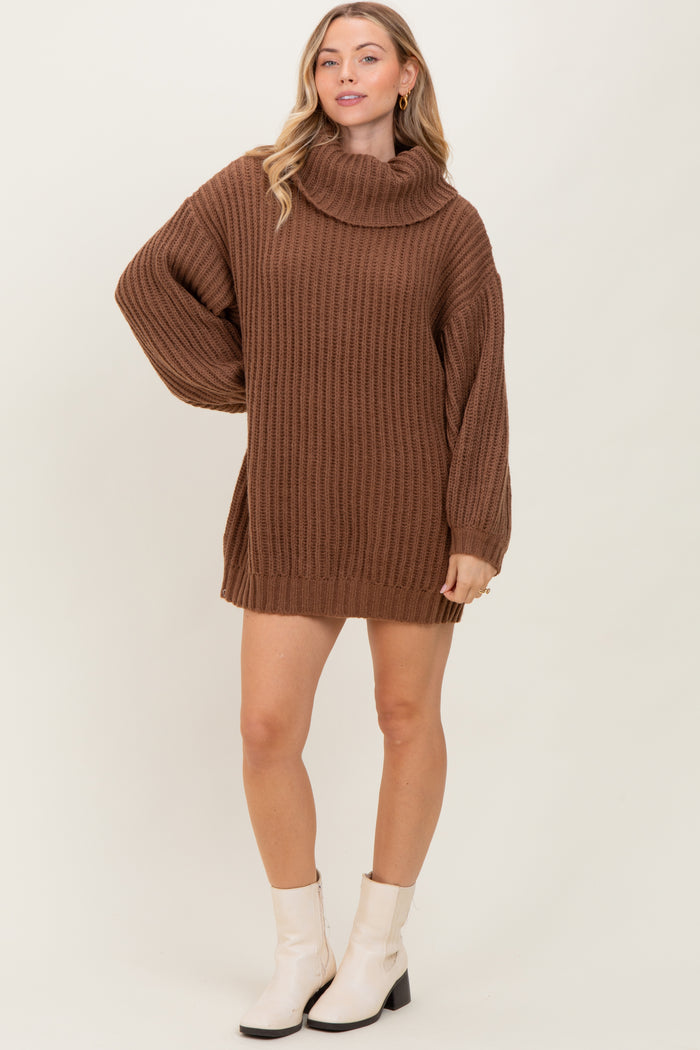 woman wearing a brown chunky knit sweater dress with a cowl neck and long sleeves, standing with one hand on her hip