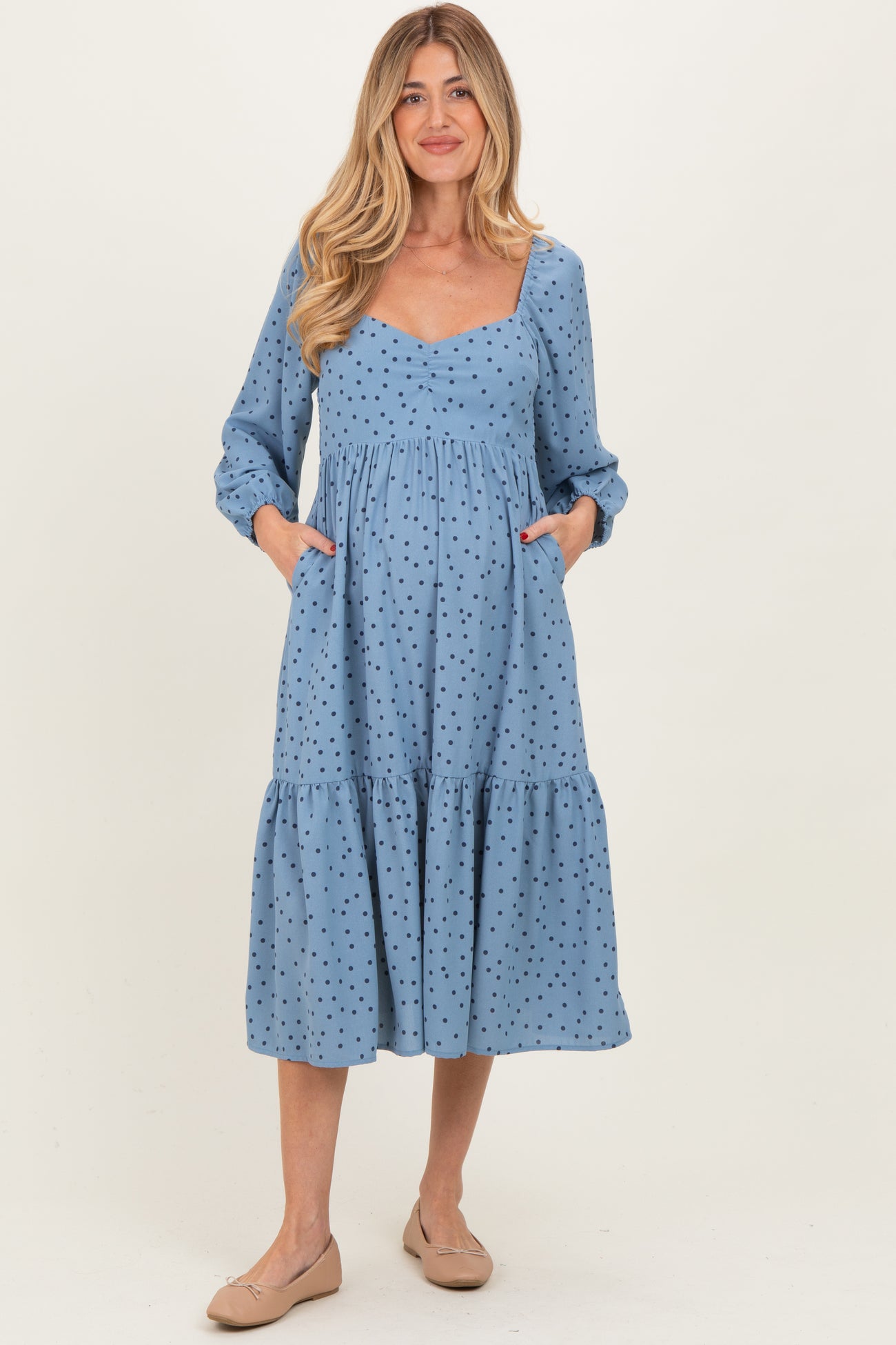 woman wearing a blue polka dot maternity maxi dress with a square neckline and pockets, showing off her baby bump