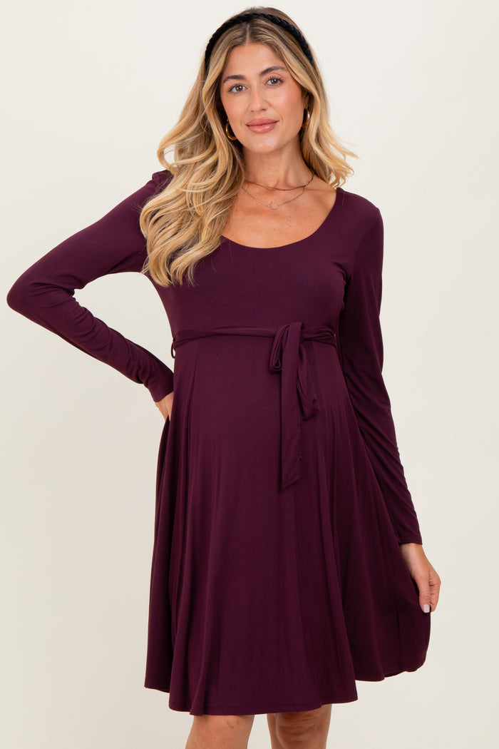 woman wearing a dark purple long sleeve maternity dress with a tie waist and a visible baby bump