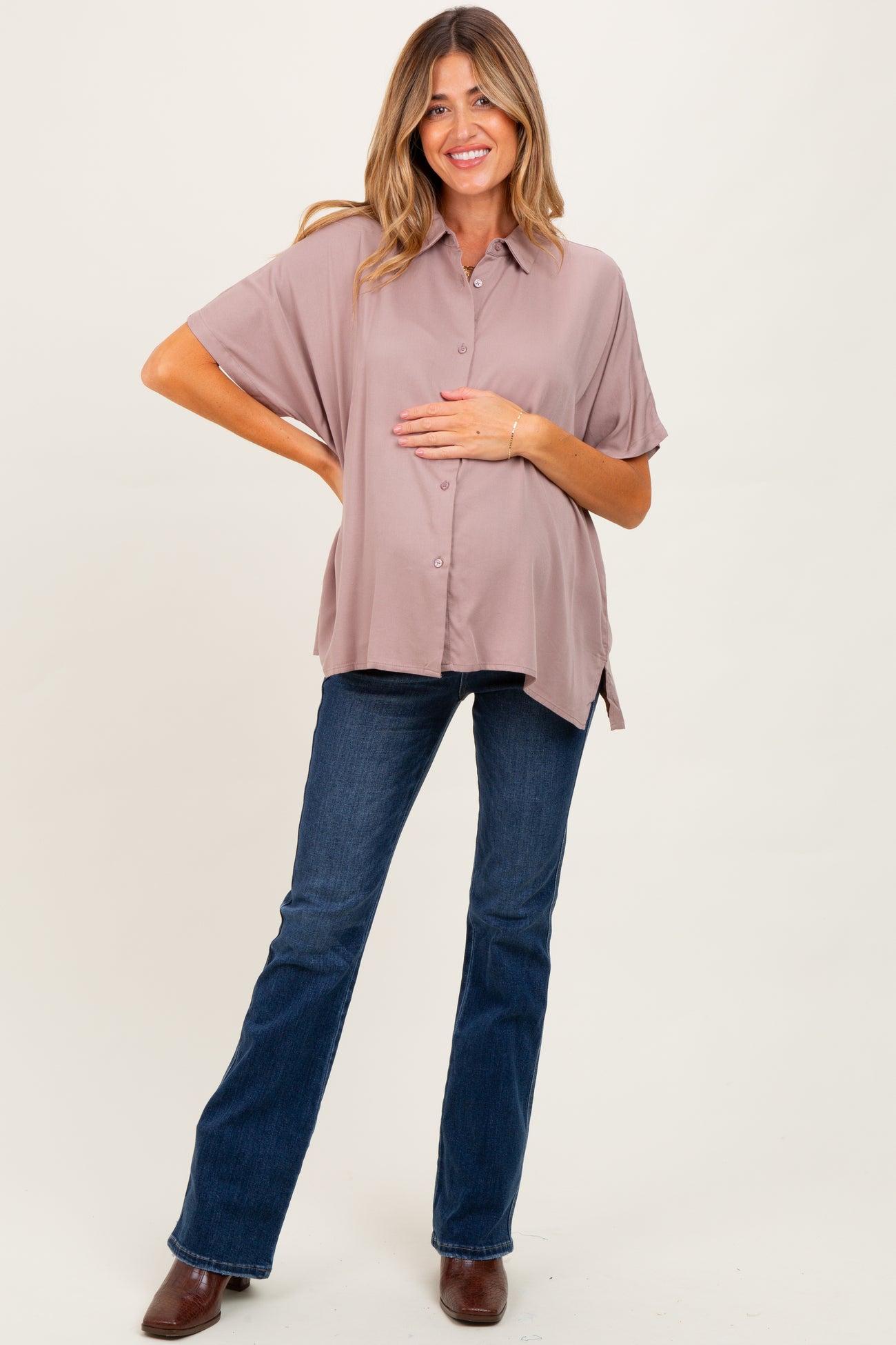 woman wearing a mauve short sleeve maternity button-up shirt and dark blue flared jeans, gently holding her baby bump