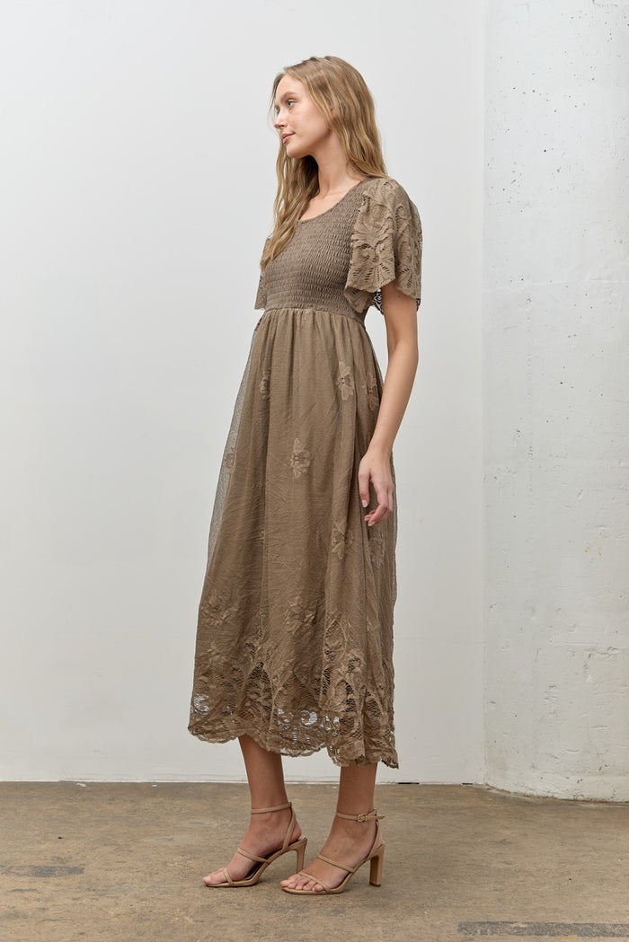 woman wearing a brown maxi dress with embroidered details and short sleeves, standing sideways against a white wall