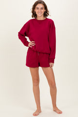 woman wearing a burgundy oversized sweatshirt and matching shorts, standing barefoot against a neutral background