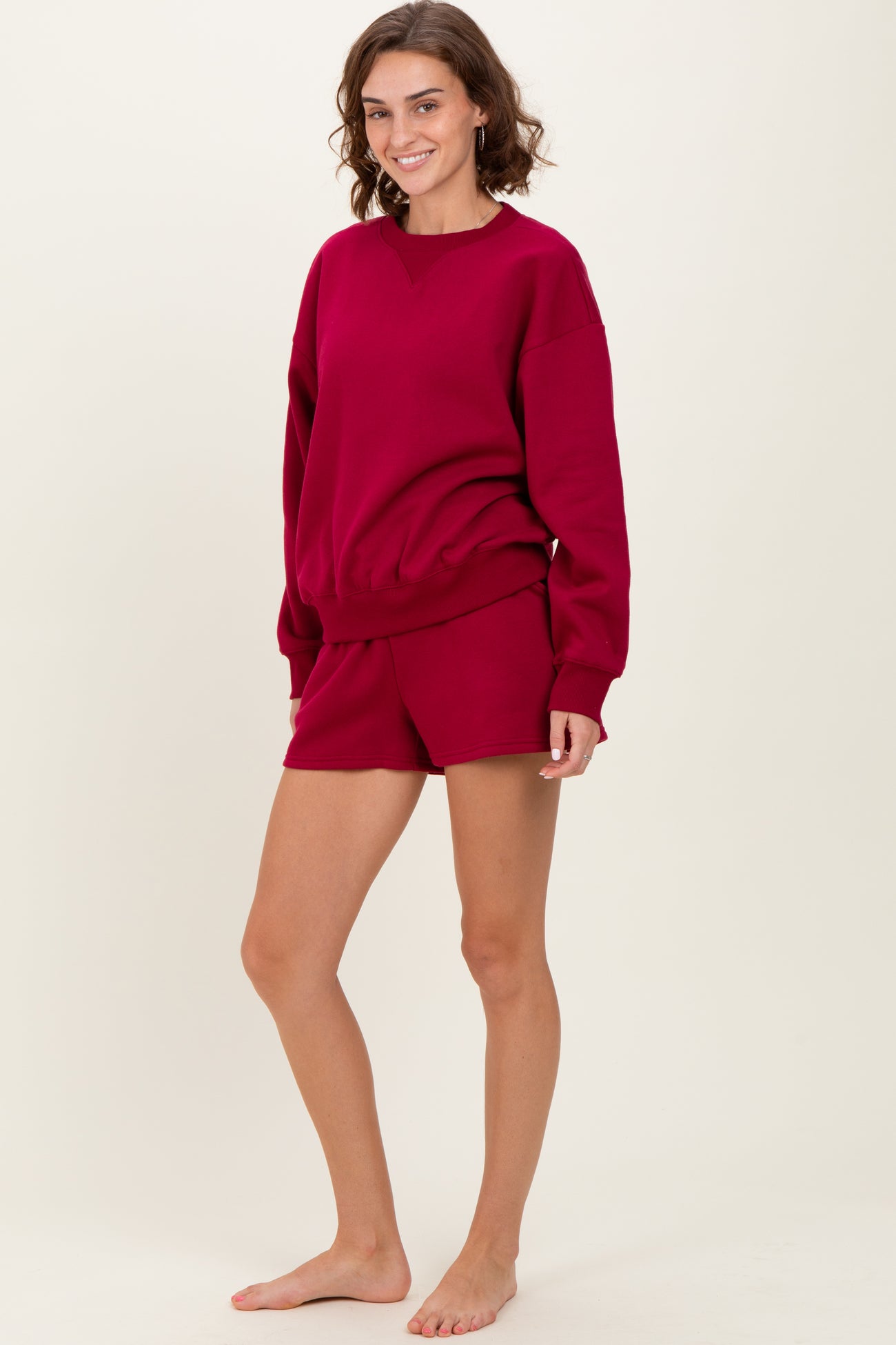 woman wearing a burgundy oversized sweatshirt and matching shorts, standing barefoot on a neutral background