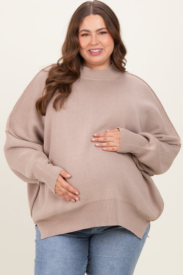 woman wearing a beige oversized maternity sweater and holding her baby bump, paired with blue jeans
