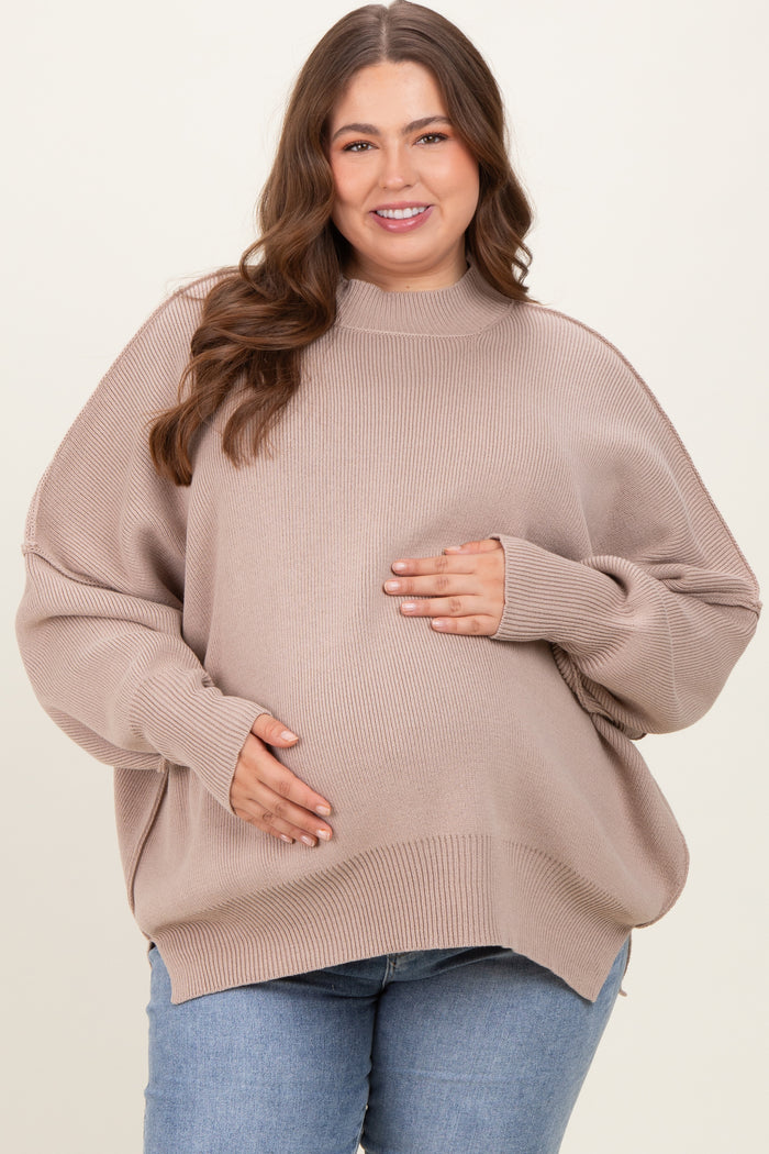 woman wearing a beige oversized maternity sweater and holding her baby bump, paired with blue jeans
