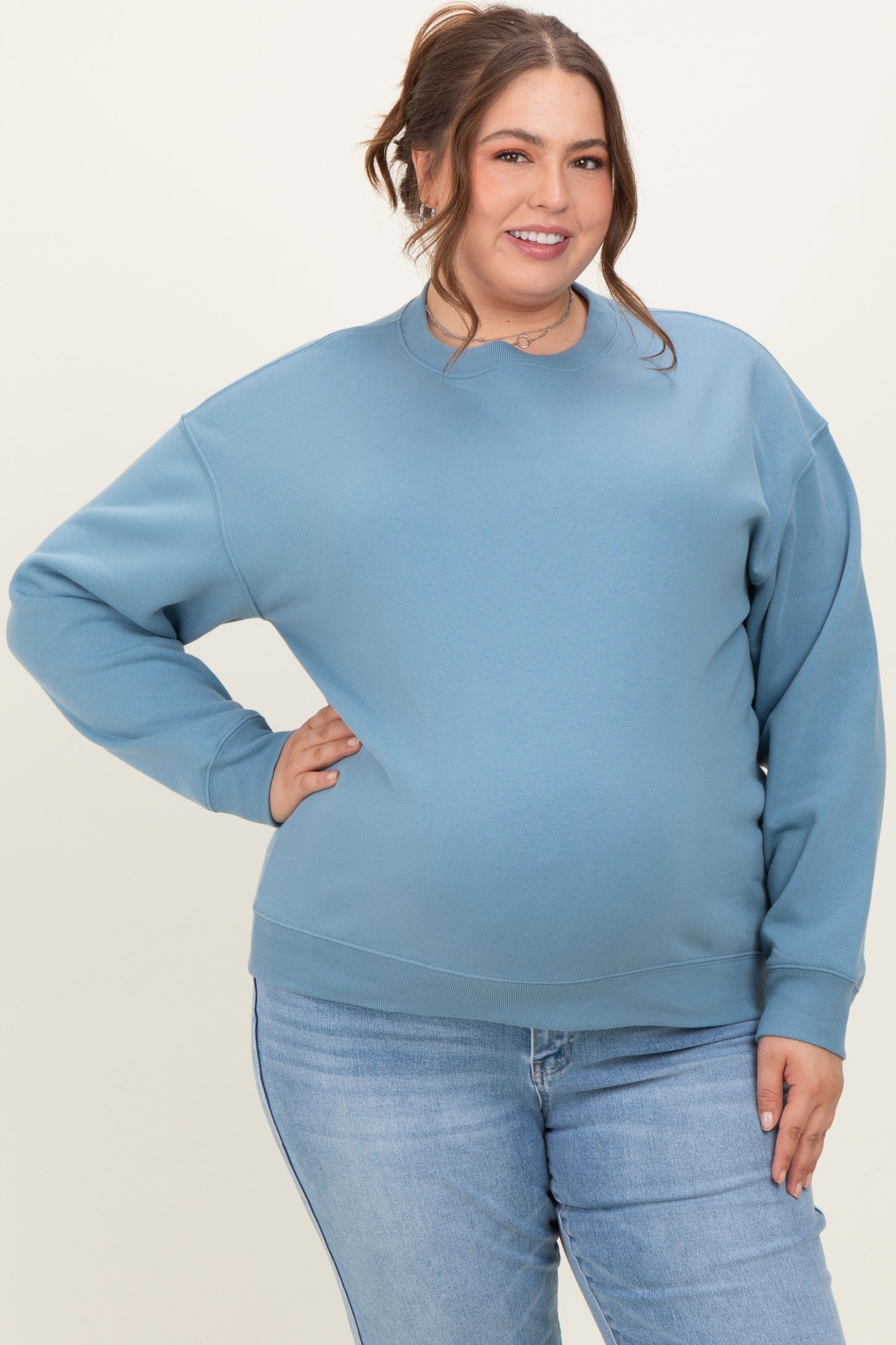 woman wearing a light blue oversized maternity sweatshirt and showing her baby bump with a smile while posing