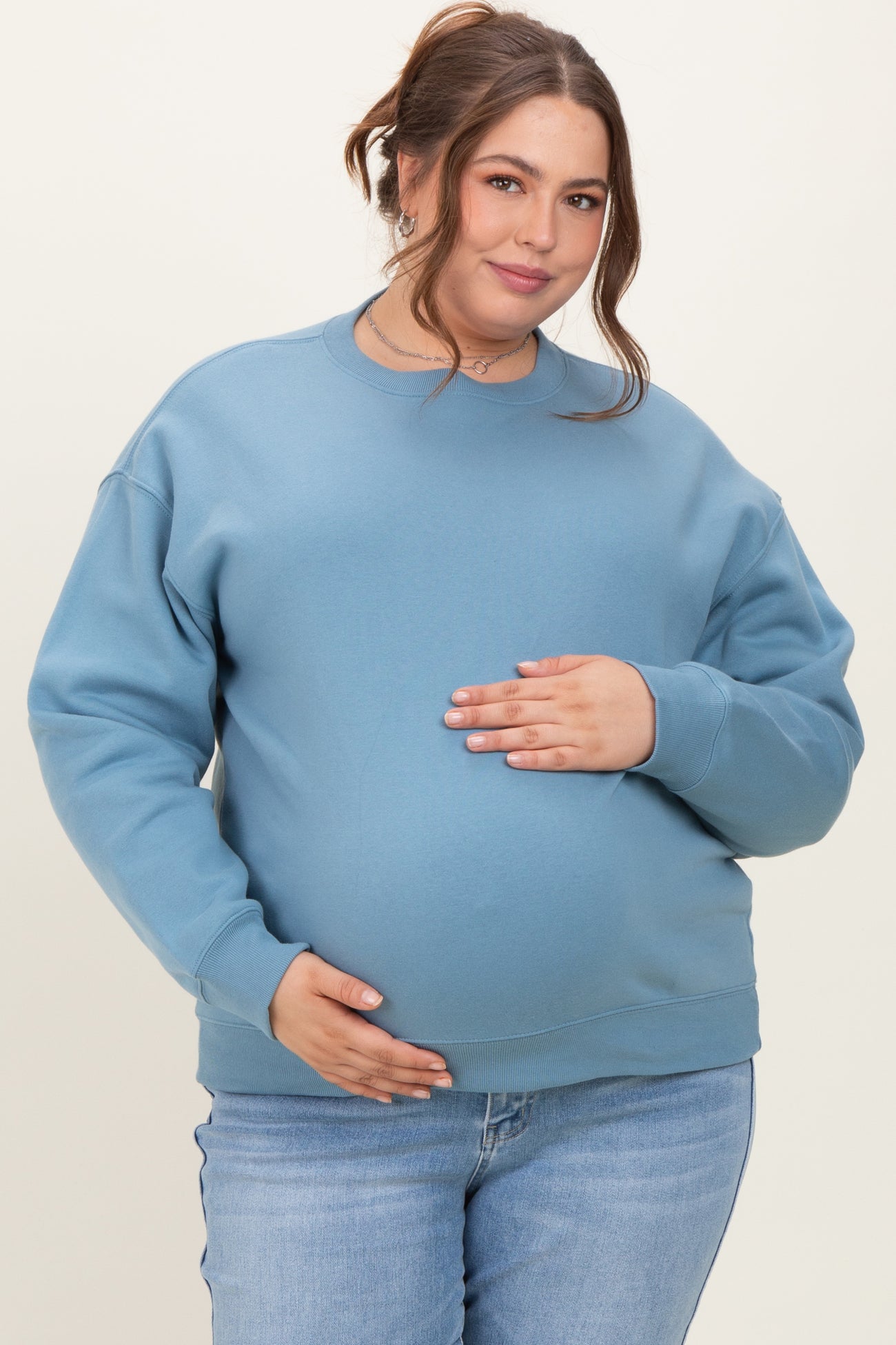 woman wearing a blue maternity sweatshirt, holding her baby bump, paired with light denim jeans