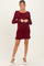 woman wearing a burgundy maternity dress with ruffled sleeves and hem, posing with hands clasped at her waist