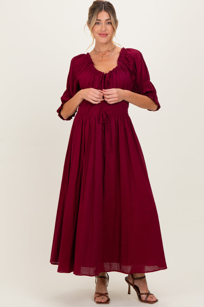 woman wearing a burgundy maternity maxi dress with puff sleeves and a drawstring waist, holding the fabric at the front