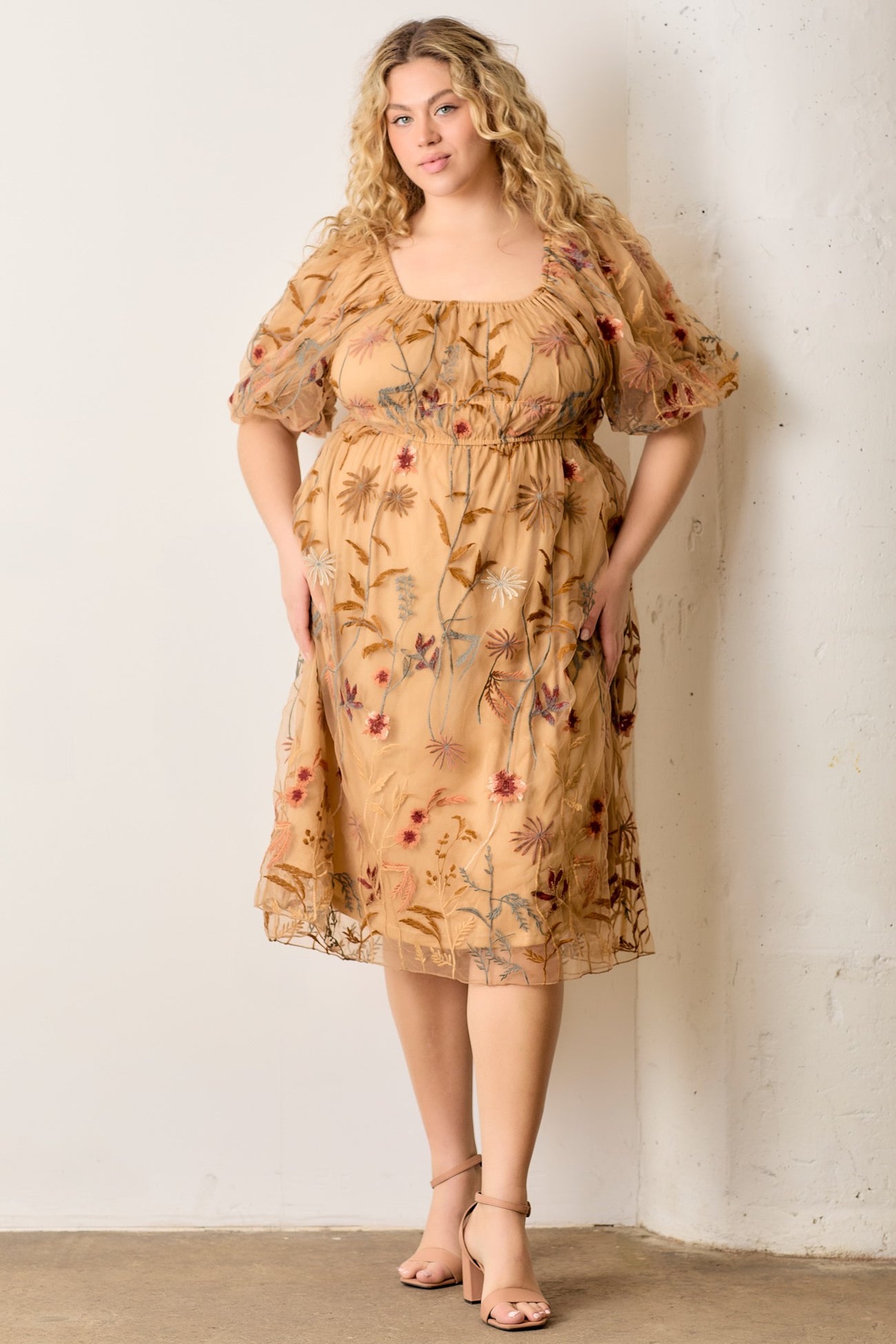 woman wearing a beige plus-size dress with floral embroidery and puff sleeves, standing against a wall