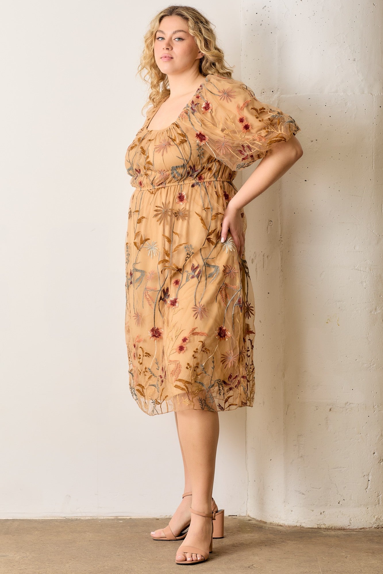 woman wearing a floral embroidered plus size dress with puff sleeves and a square neckline, standing against a wall