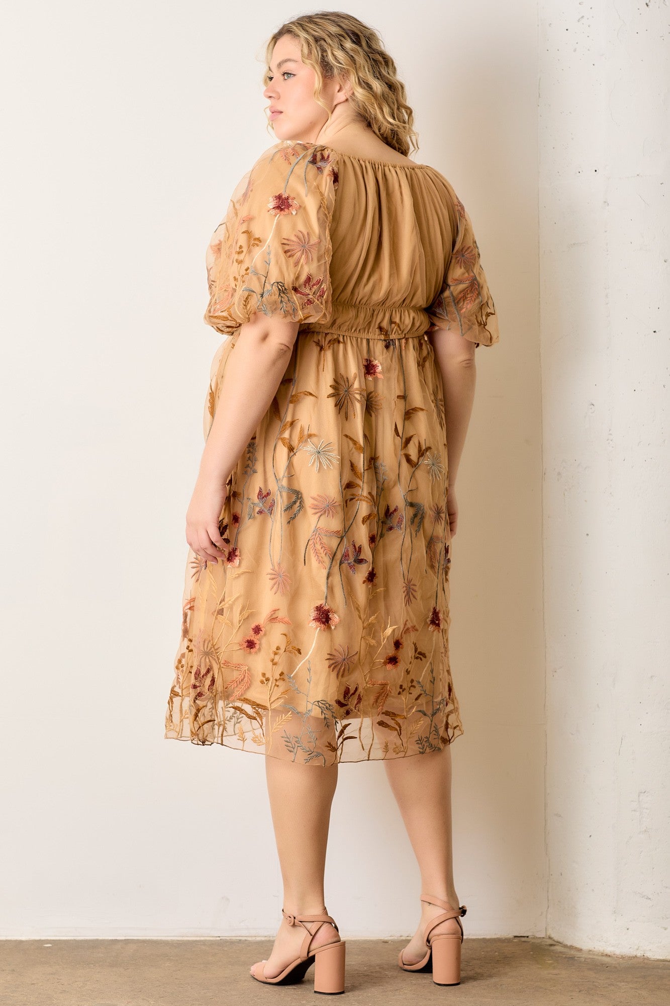 woman wearing a beige plus size dress with floral embroidery and puff sleeves, standing against a white wall