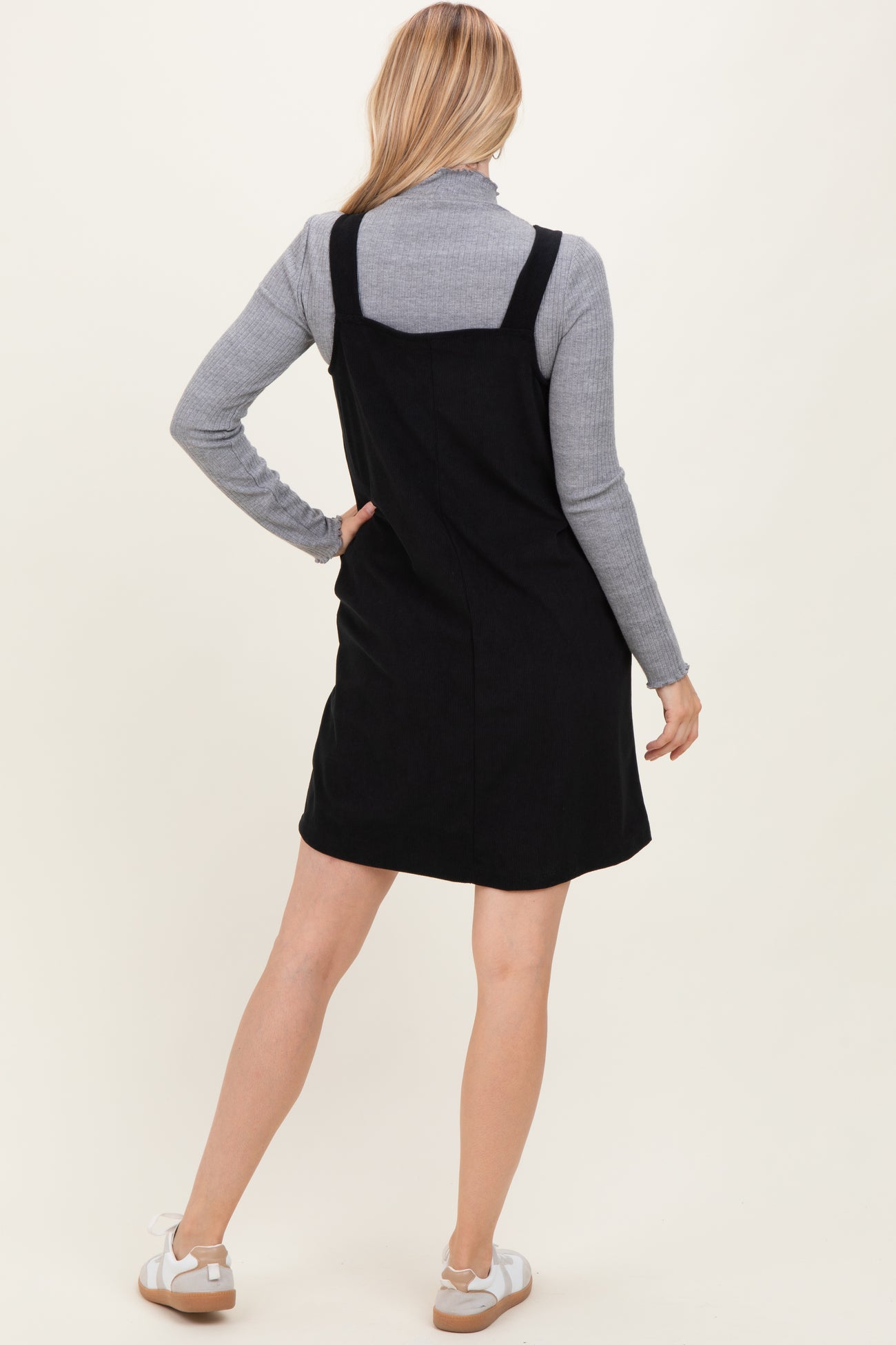 woman wearing a black denim overall dress over a grey turtleneck sweater, viewed from the back