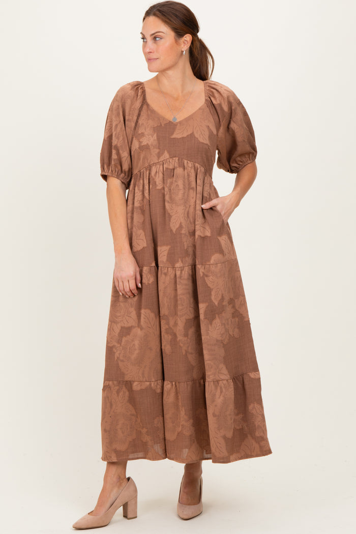 woman wearing a brown floral maxi dress with puff sleeves and a tiered skirt, standing with one hand in the pocket