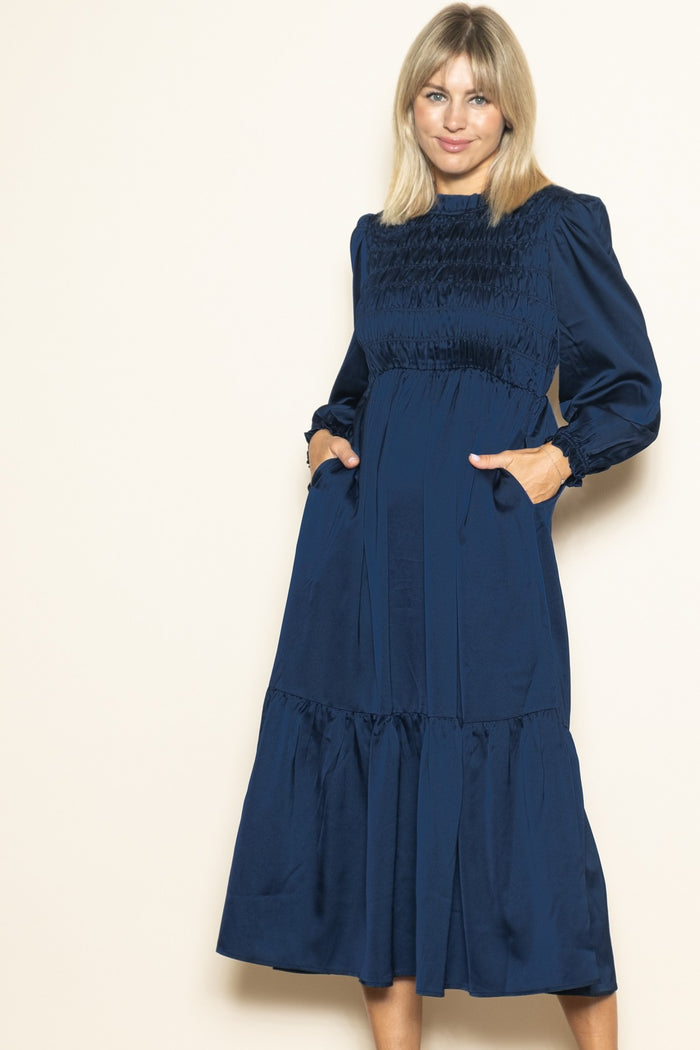 woman wearing a navy blue long sleeve dress with smocked bodice and pockets, standing with a smile