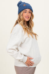 woman wearing a light gray maternity sweatshirt and a blue knitted beanie, cradling her baby bump