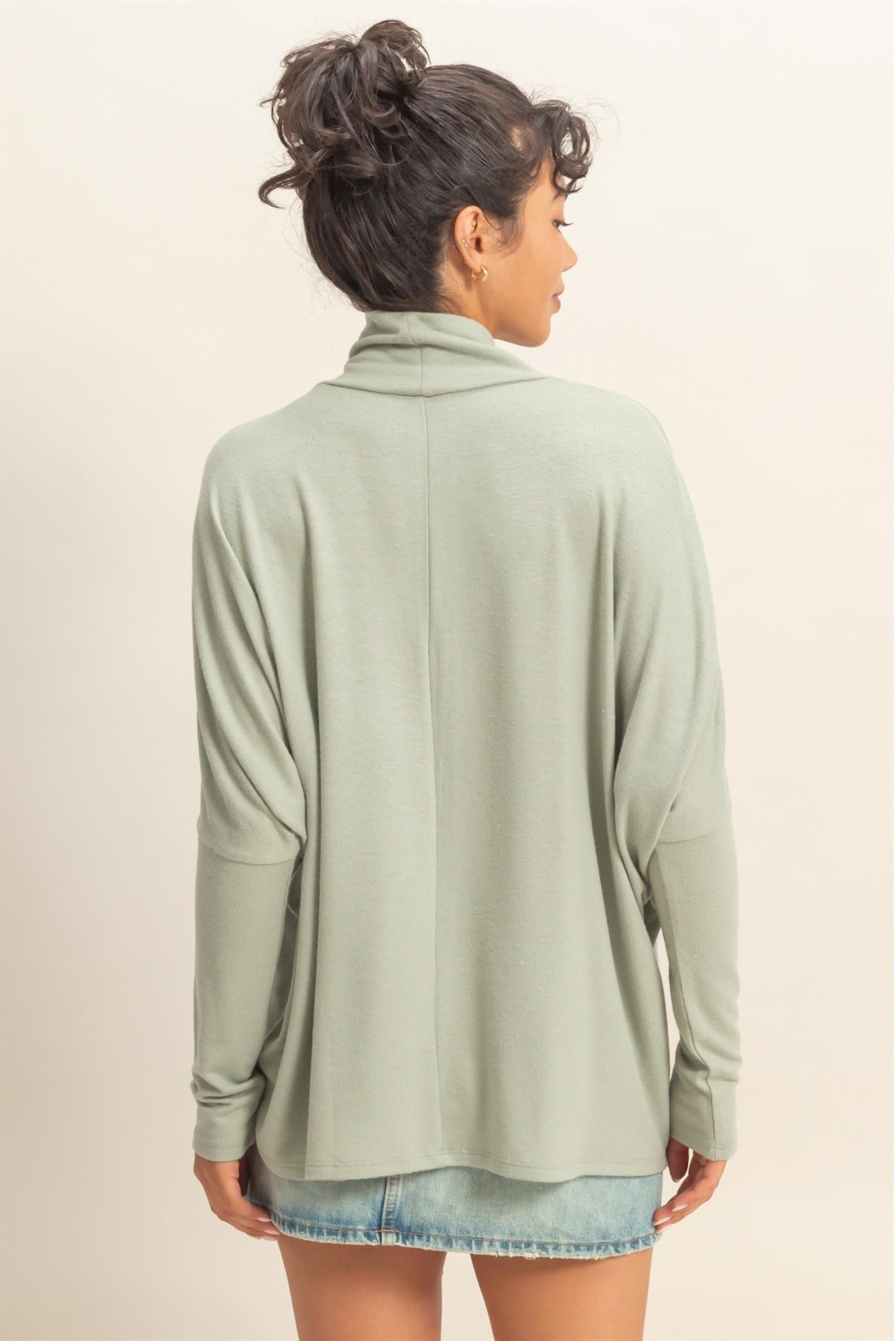 woman wearing a light green long sleeve pullover with a relaxed fit and back drape detail, styled with a denim mini skirt