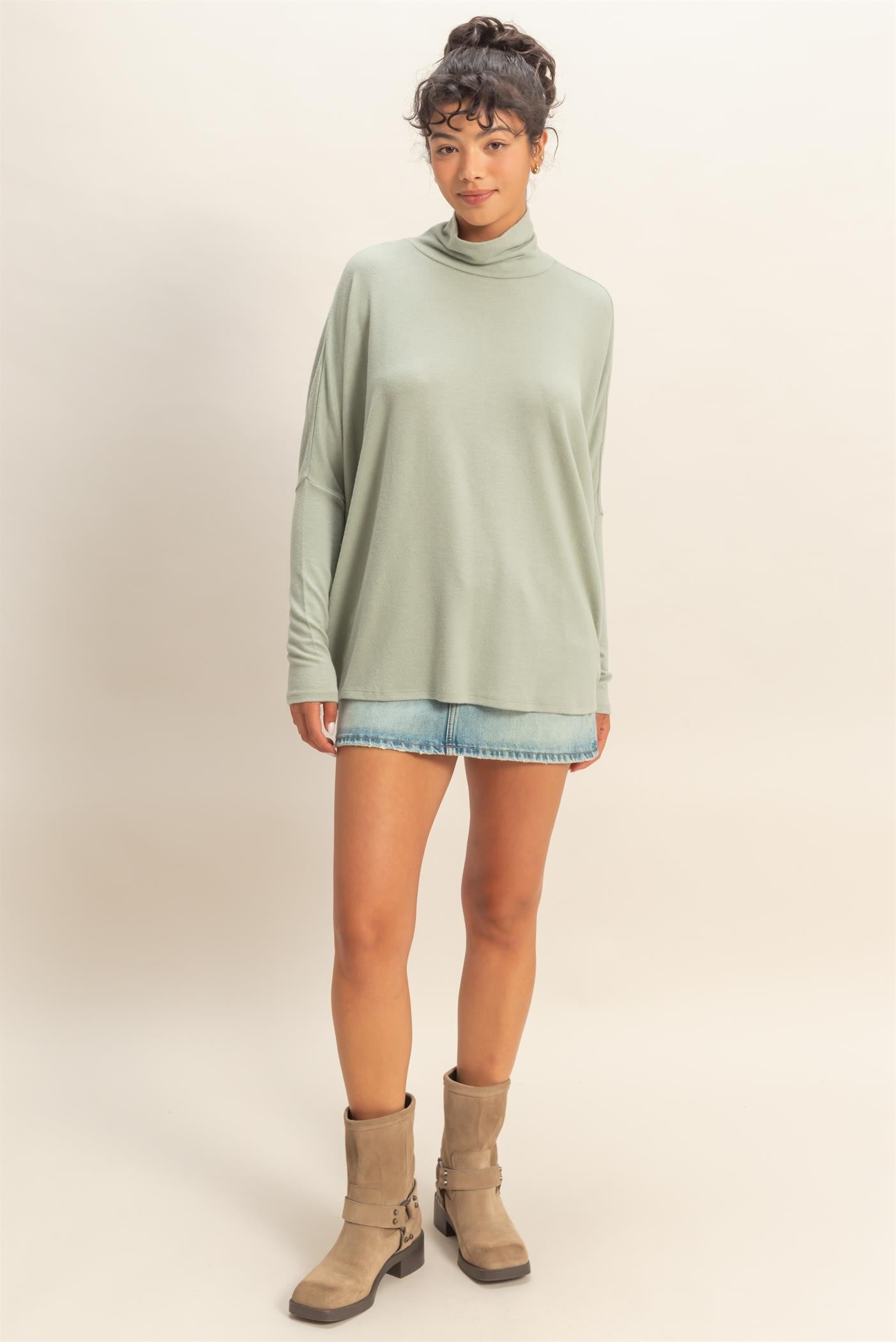 woman wearing a light green long sleeve mock neck top with loose fit and denim shorts, paired with beige ankle boots
