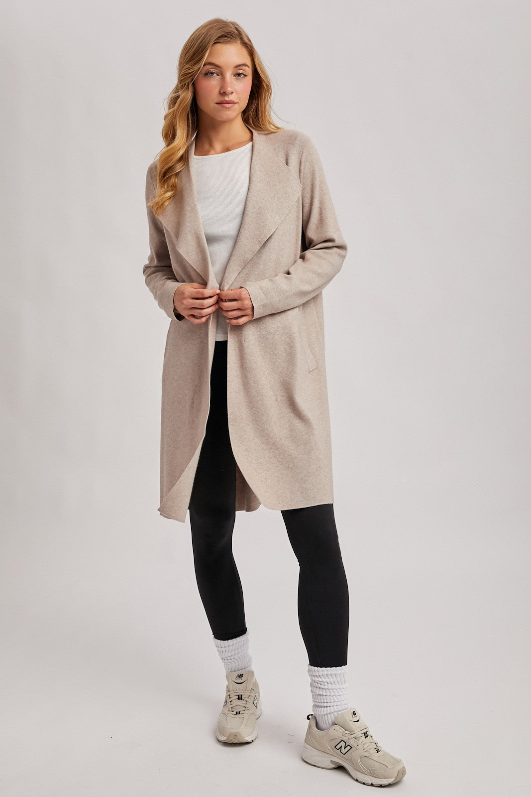 woman wearing a light beige long cardigan over a white top and black leggings, paired with white socks and sneakers