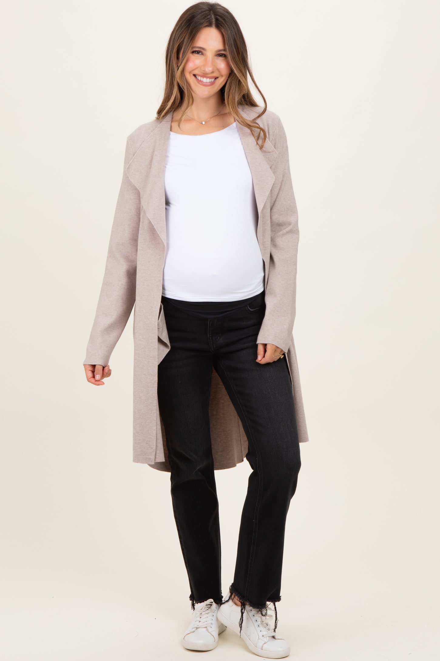woman wearing a light pink maternity long cardigan over a white top and black jeans, showing her baby bump