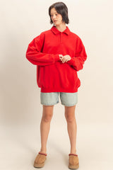 woman wearing a red oversized polo sweatshirt with long sleeves and denim shorts, standing in casual footwear