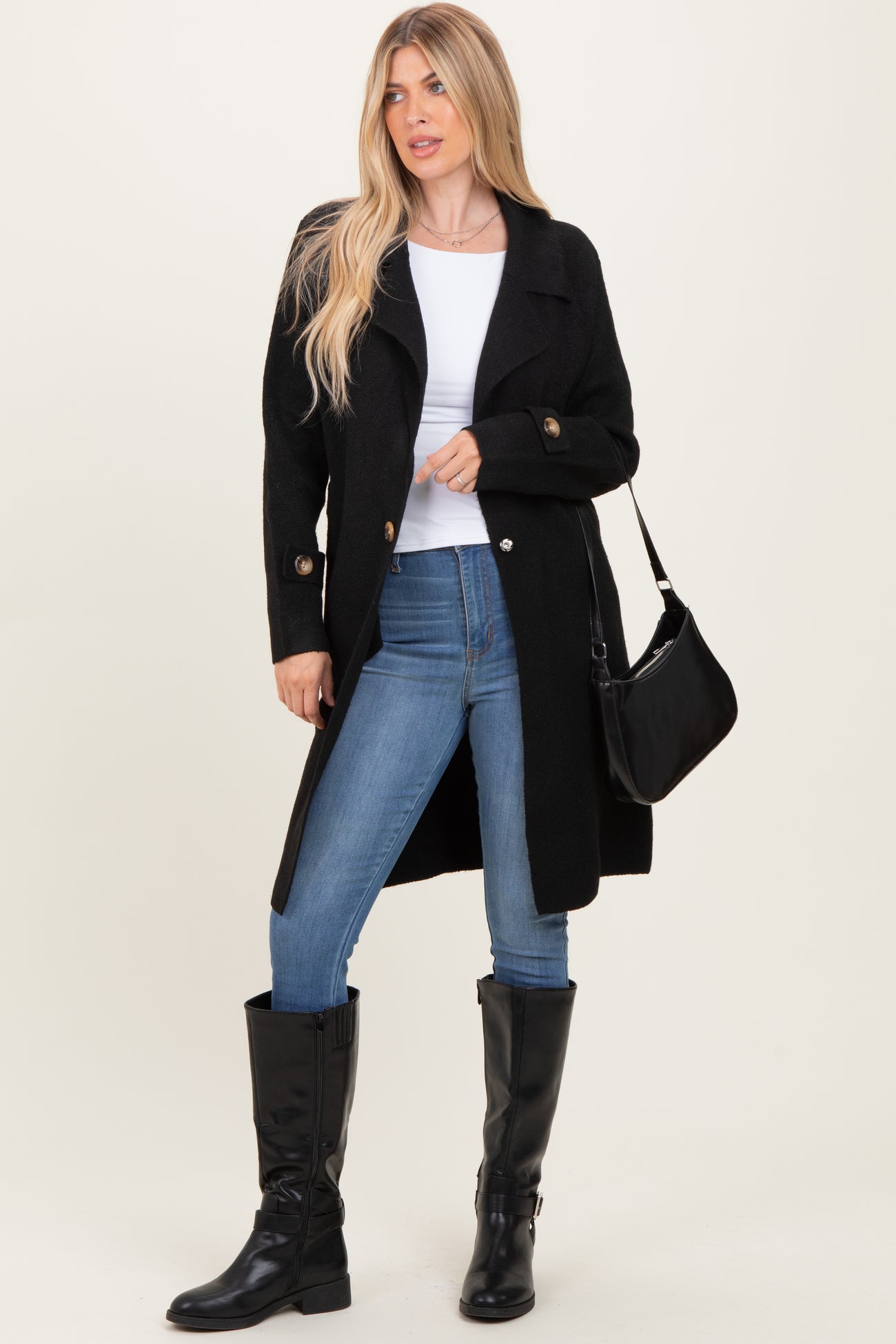 woman wearing a black long coat over a white top, paired with jeans and knee-high black boots, holding a black bag