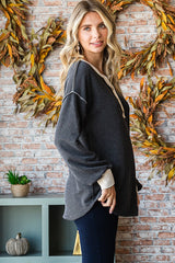 woman wearing a gray ribbed oversized sweater with a v-neck and contrasting cream cuffs, standing in front of decorative fall foliage