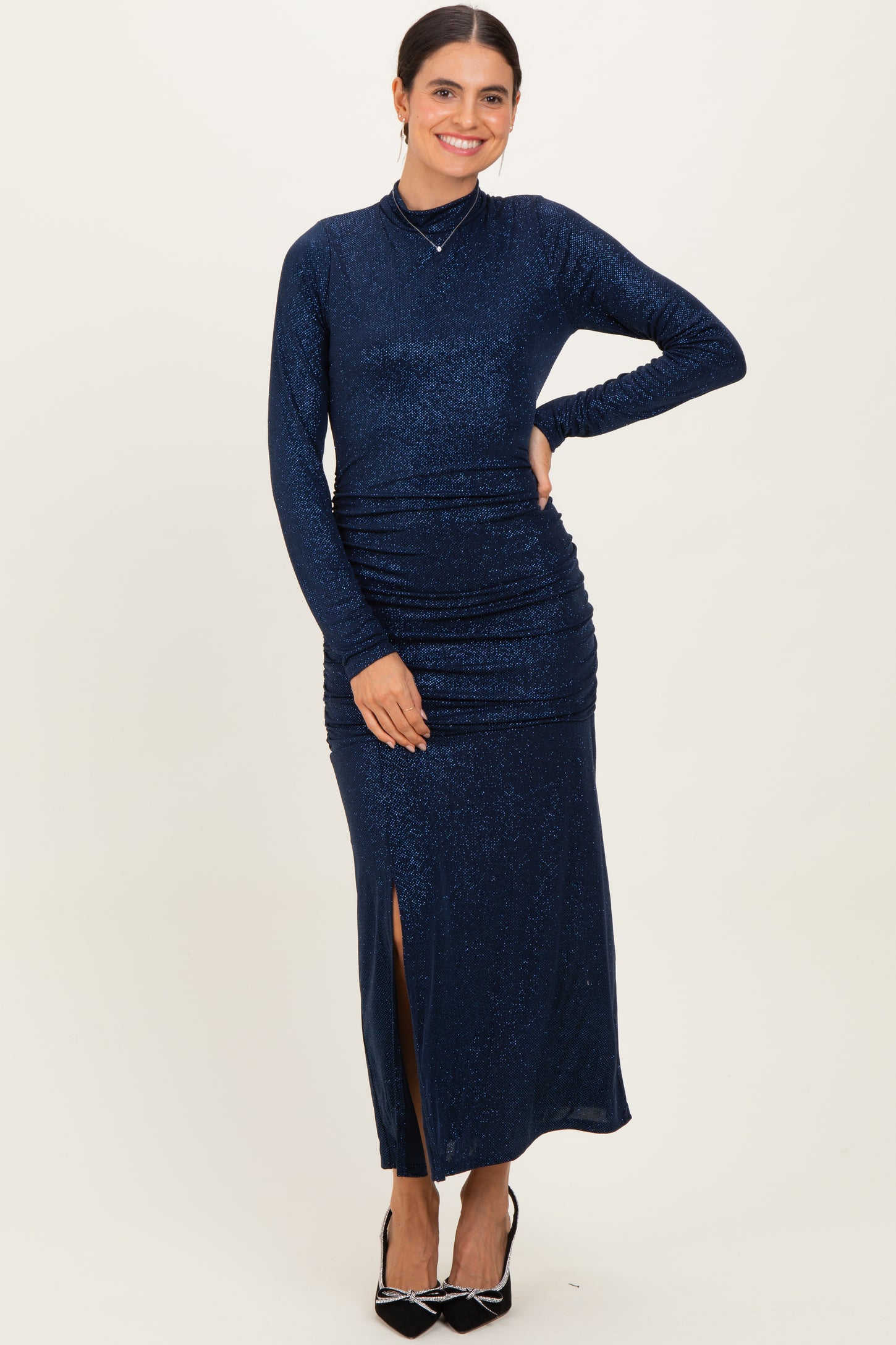 woman wearing a navy blue long sleeve shimmer maxi dress with ruching and a side slit, posing with one hand on her hip