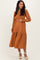 woman wearing a rust colored maternity midi dress with long sleeves and a smocked bodice, showing her baby bump