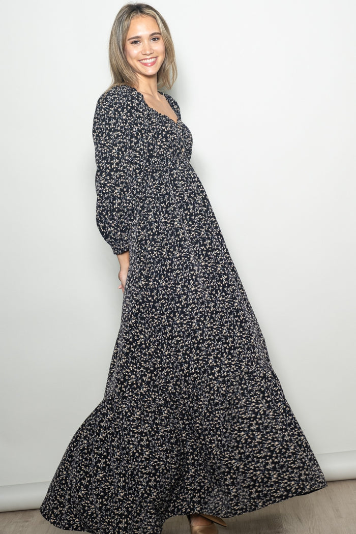 woman wearing a black floral maxi dress with long sleeves and a fitted bodice, smiling against a neutral background