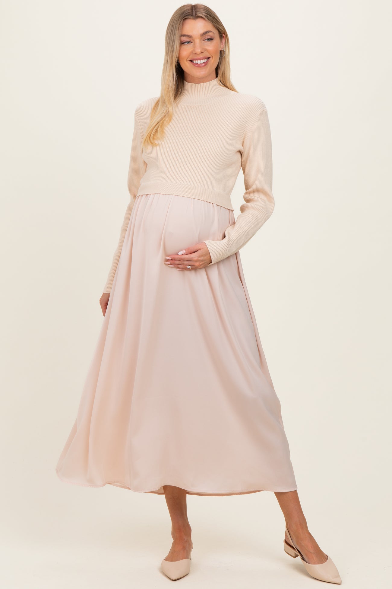 woman wearing a cream ribbed maternity sweater and a flowing blush pink maternity maxi skirt, holding her baby bump