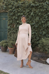 woman wearing a beige maternity sweater and a flowy beige maternity dress, holding a brown handbag and standing outdoors