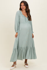 woman wearing a sage green long sleeve maternity dress with button details and a tiered skirt, showcasing her baby bump