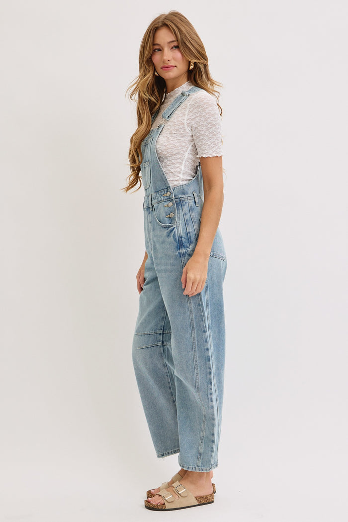 woman wearing light blue denim overalls over a white polka dot top, standing sideways with long hair and beige sandals