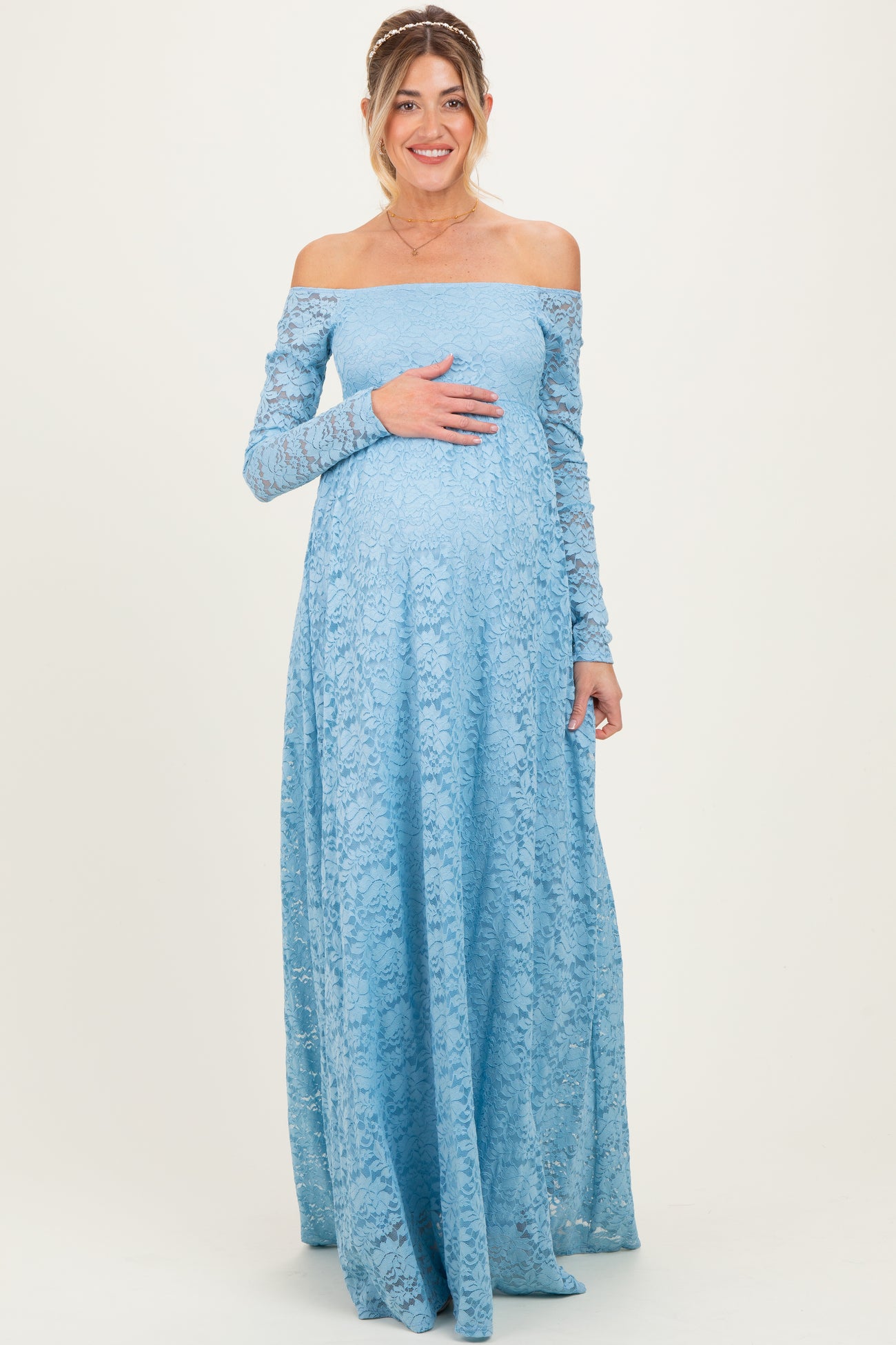 woman wearing a light blue off-the-shoulder lace maternity gown with long sleeves and cradling her baby bump