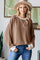 woman wearing a brown ribbed oversized sweater with white cuffs and a wide-brimmed hat, standing in a stylish interior