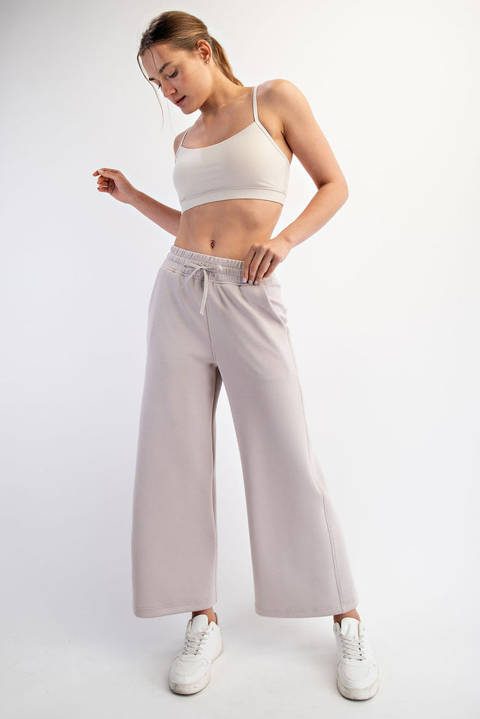 woman wearing a beige cropped tank top and light gray wide-leg pants, standing in a casual pose with white sneakers