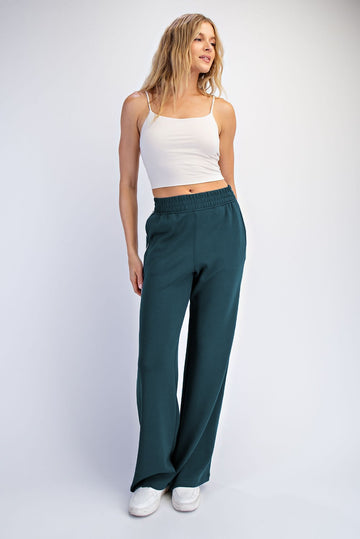 woman wearing a white cropped tank top and teal wide-leg pants, standing with one hand on her hip